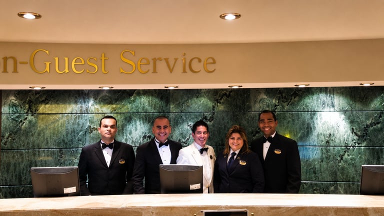 Five hotel staff members in formal black uniforms standing behind a front desk with a Guest Service sign