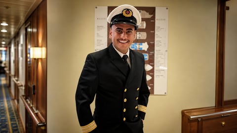 Man in naval officer uniform with white cap and black jacket standing in a ship corridor