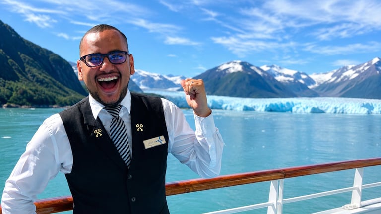 Smiling cruise ship crew member in uniform standing at ship railing with glacier and snow-capped mountains across turquoise water
